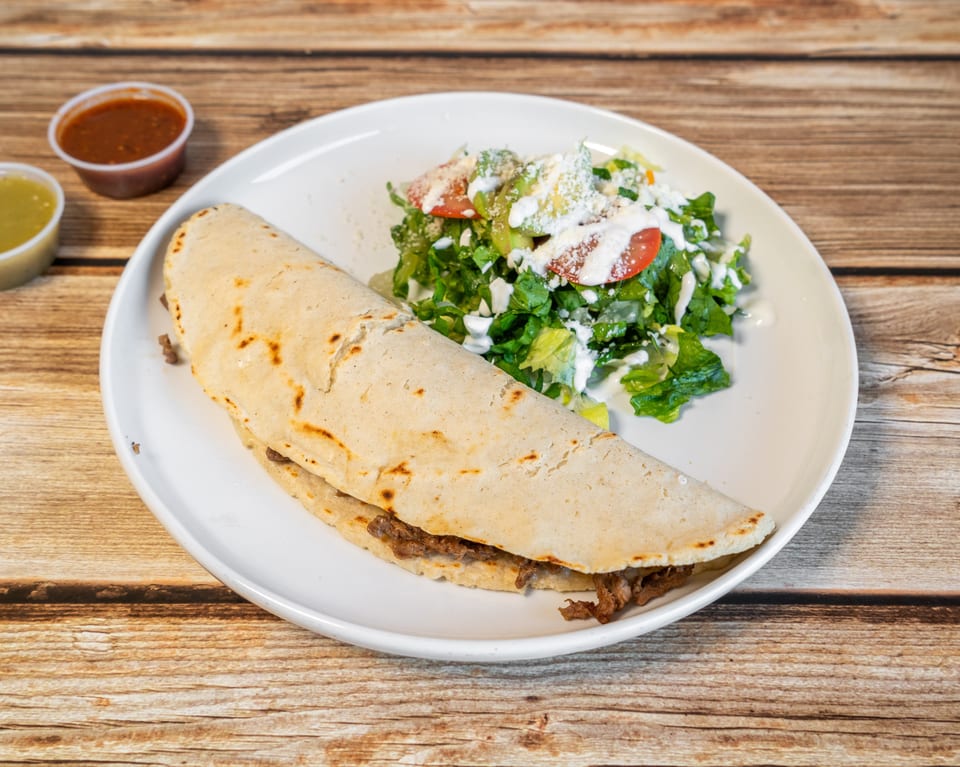 A large quesadilla with meat, a side salad with dressing, and two small sauce cups on a wooden table.