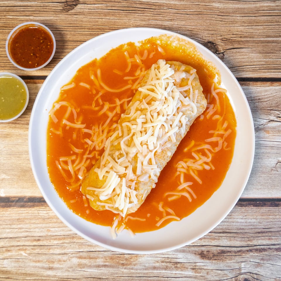 A cheese-covered burrito drenched in red sauce, with green and red salsas on a wooden table.