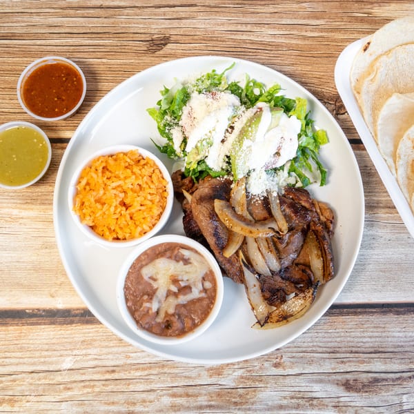 Carne asada with grilled onions, rice, beans, salad, tortillas, and two salsas on a wooden table.