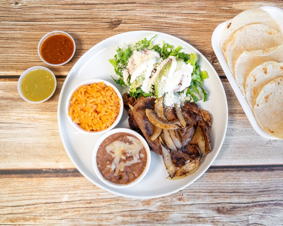 Carne asada with grilled onions, rice, beans, salad, tortillas, and two salsas on a wooden table.