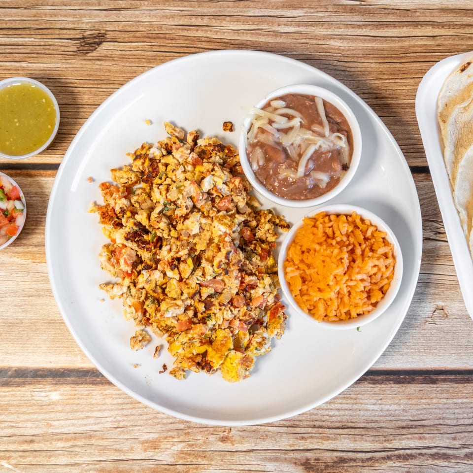 Scrambled eggs with pico de gallo, beans, rice, salsa verde, and tortillas on a wooden table.