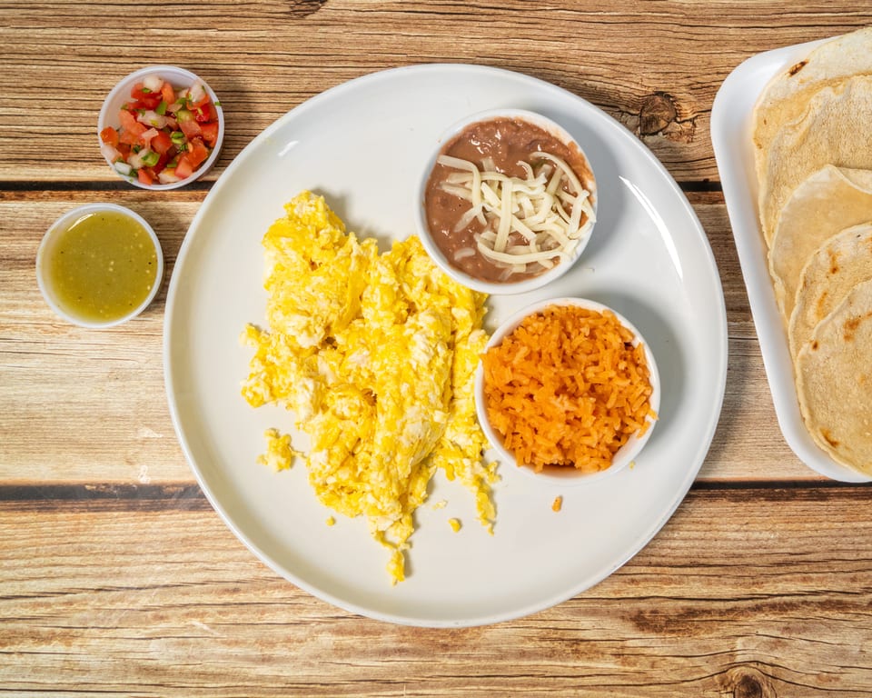 Scrambled eggs, refried beans with cheese, rice, salsa, pico de gallo, and tortillas on a wooden table.