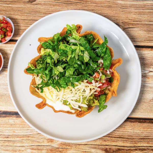 Taco salad in a fried tortilla bowl with lettuce, cheese, guacamole, and salsa on a white plate.