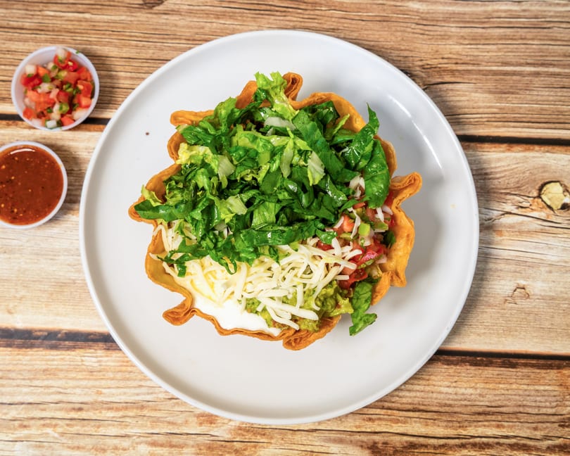 Taco salad in a fried tortilla bowl with lettuce, cheese, guacamole, and salsa on a white plate.