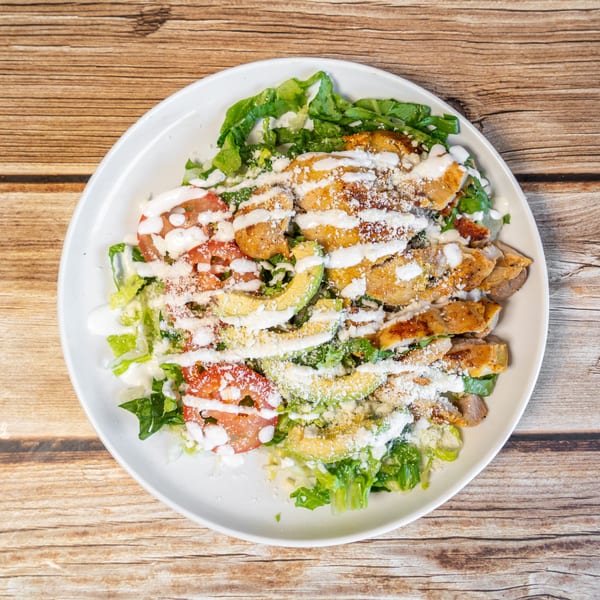 Grilled chicken salad with lettuce, tomatoes, avocado, cheese, and white dressing on a wooden table.
