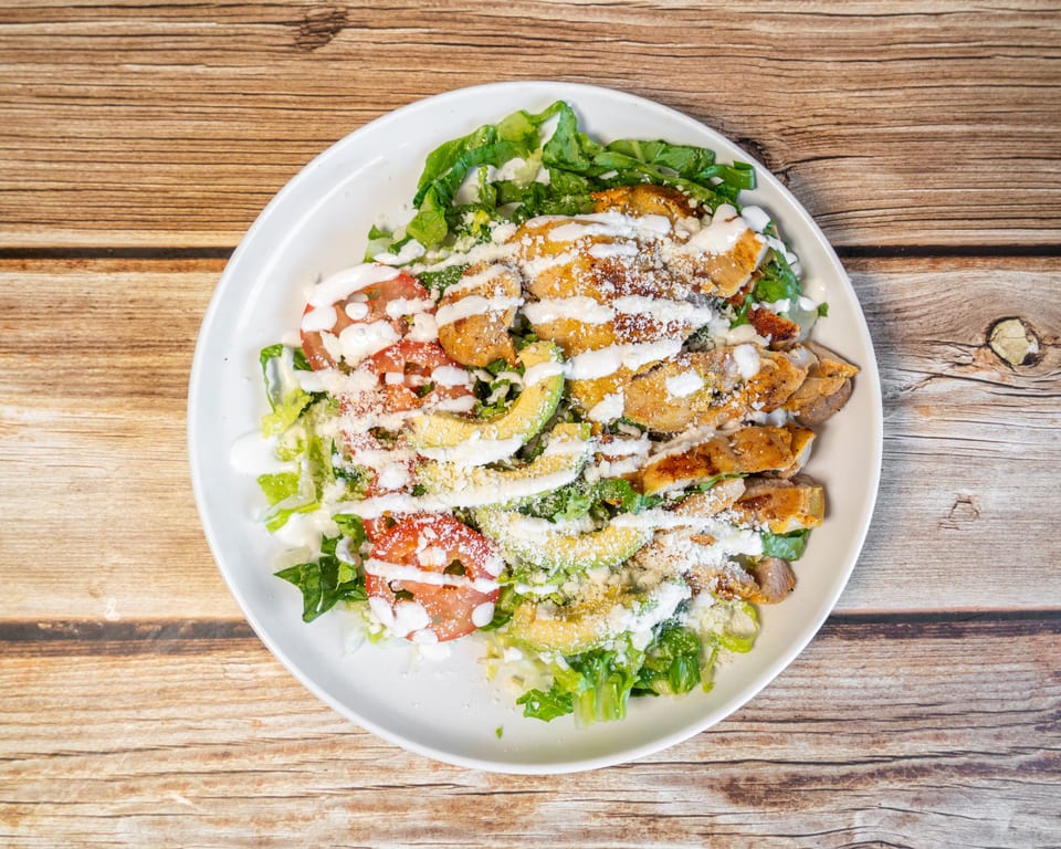 Grilled chicken salad with lettuce, tomatoes, avocado, cheese, and white dressing on a wooden table.
