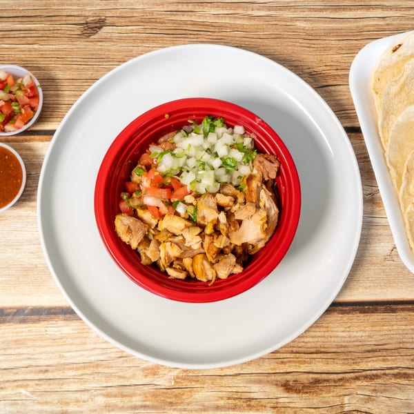A red bowl of chopped chicken, diced onions, and tomatoes, with tortillas and salsa on a wooden table.