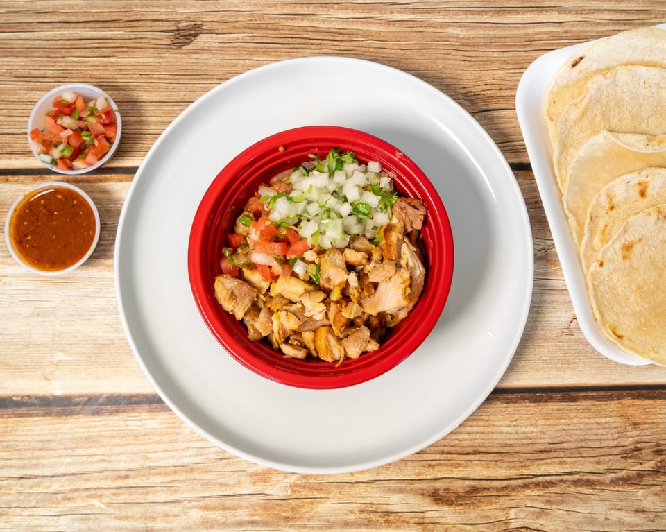 A red bowl of chopped chicken, diced onions, and tomatoes, with tortillas and salsa on a wooden table.
