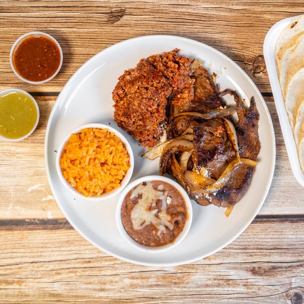 Grilled steak with onions, chorizo, rice, refried beans, two salsas, and tortillas on a wooden table.