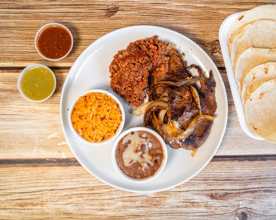 Grilled steak with onions, chorizo, rice, refried beans, two salsas, and tortillas on a wooden table.