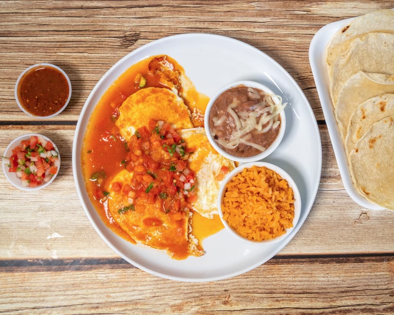 Huevos rancheros with rice, beans, salsa, and tortillas on a wooden table.