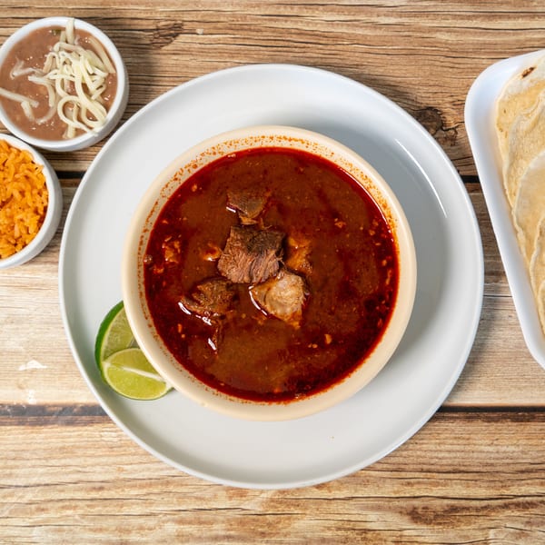 Hearty red beef stew with lime, rice, refried beans with cheese, and tortillas on a wooden table.