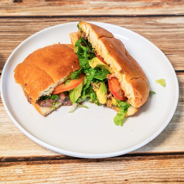 A large torta sandwich, cut in half, filled with meat, lettuce, tomato, and avocado on a white plate.