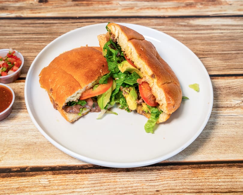 A large torta sandwich, cut in half, filled with meat, lettuce, tomato, and avocado on a white plate.