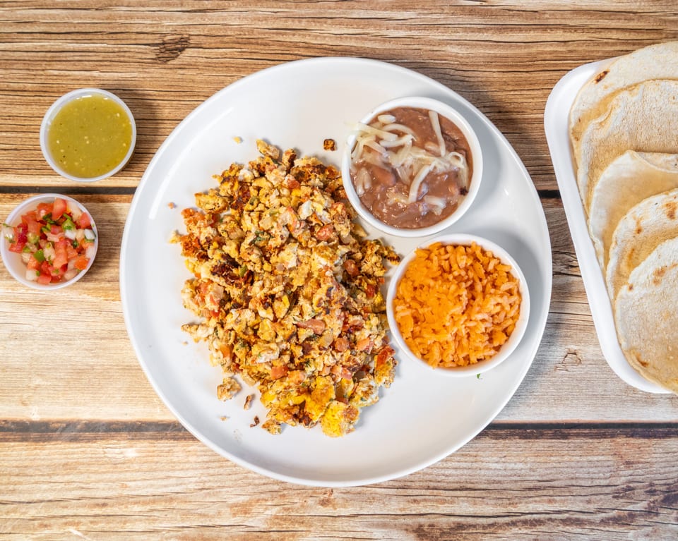Scrambled eggs with pico de gallo, rice, beans, salsa, and tortillas on a wooden table.