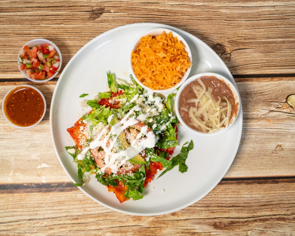 Mexican dish with enchiladas, rice, beans, salsa, and pico de gallo on a wooden table.