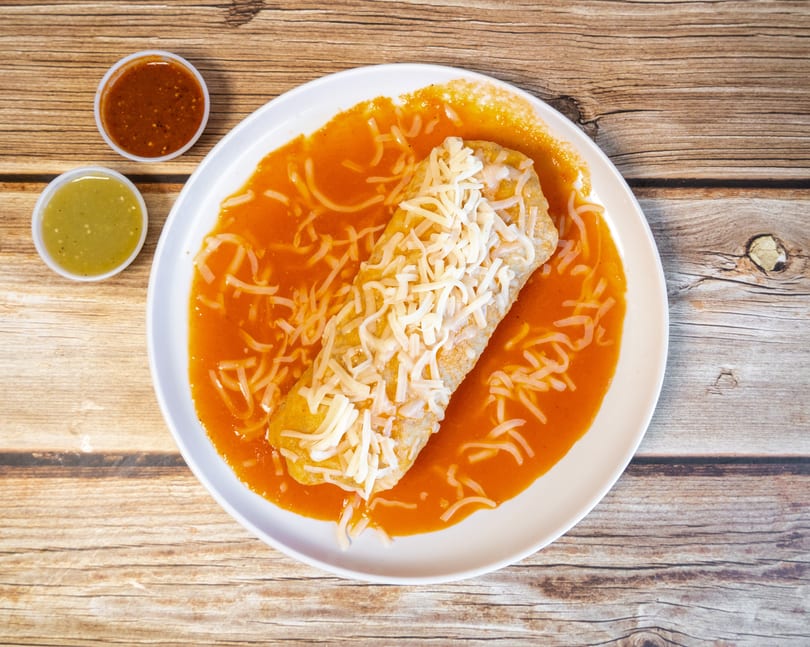 Wet burrito covered in red sauce and shredded cheese, with two small salsa cups on a wooden table.